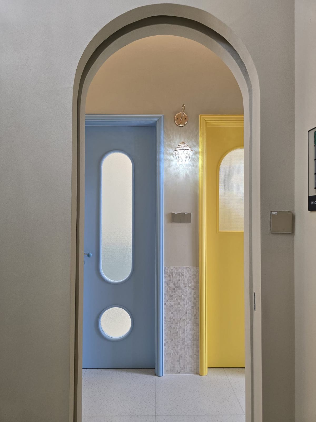 An arched opening framing a pale blue door with a pill-shaped and a round window, a yellow door with an arched glass panel, and a crystal wall light between them.