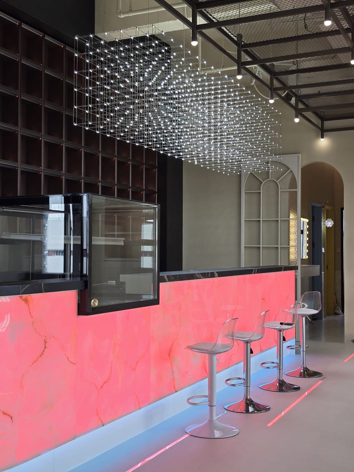 Coral backlit counter with acrylic stools under a suspended grid light installation, with a glazed arched screen at the far end.