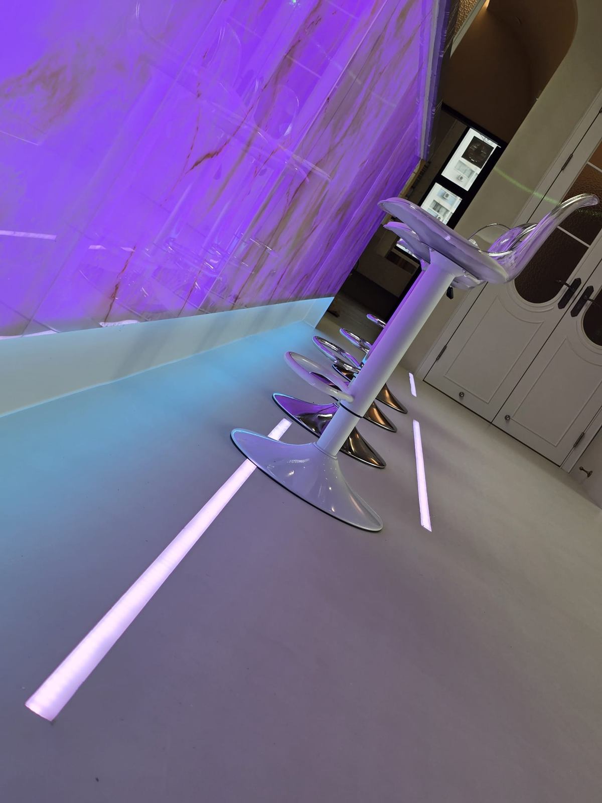 A tilted view of acrylic stools along the violet-lit counter, with an in-floor light line and white panelled doors.