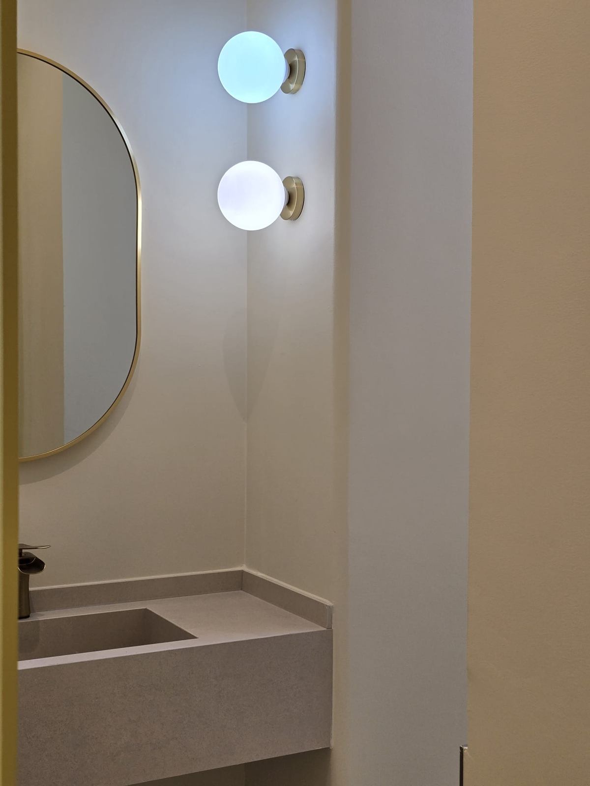 A washbasin with a stone-look basin, a brass-framed oval mirror and two glowing globe wall lights.