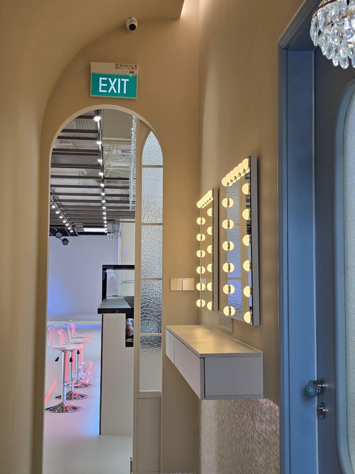 A vaulted corridor with two bulb-lit vanity mirrors over a floating white shelf, looking through an arched opening towards the studio floor.