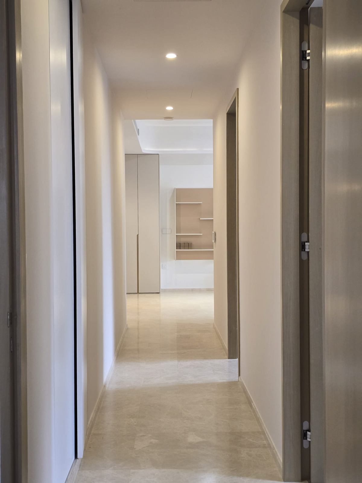 A long pale corridor with flush doors and a stone floor, ending in an open wall shelf.