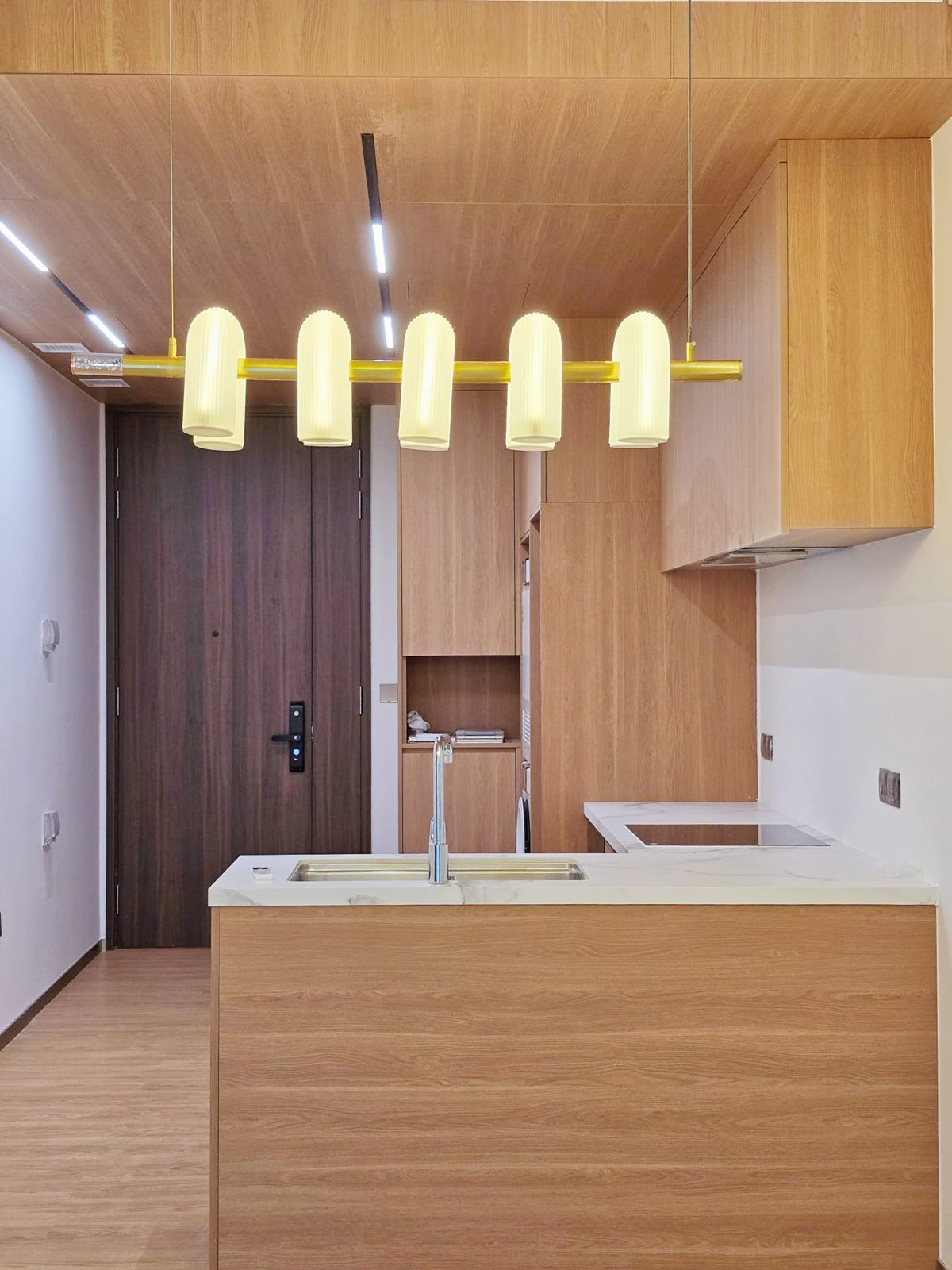 A kitchen peninsula in timber-toned veneer with a white marble-look top, under a pendant of ribbed white shades on a brass bar and a timber ceiling.