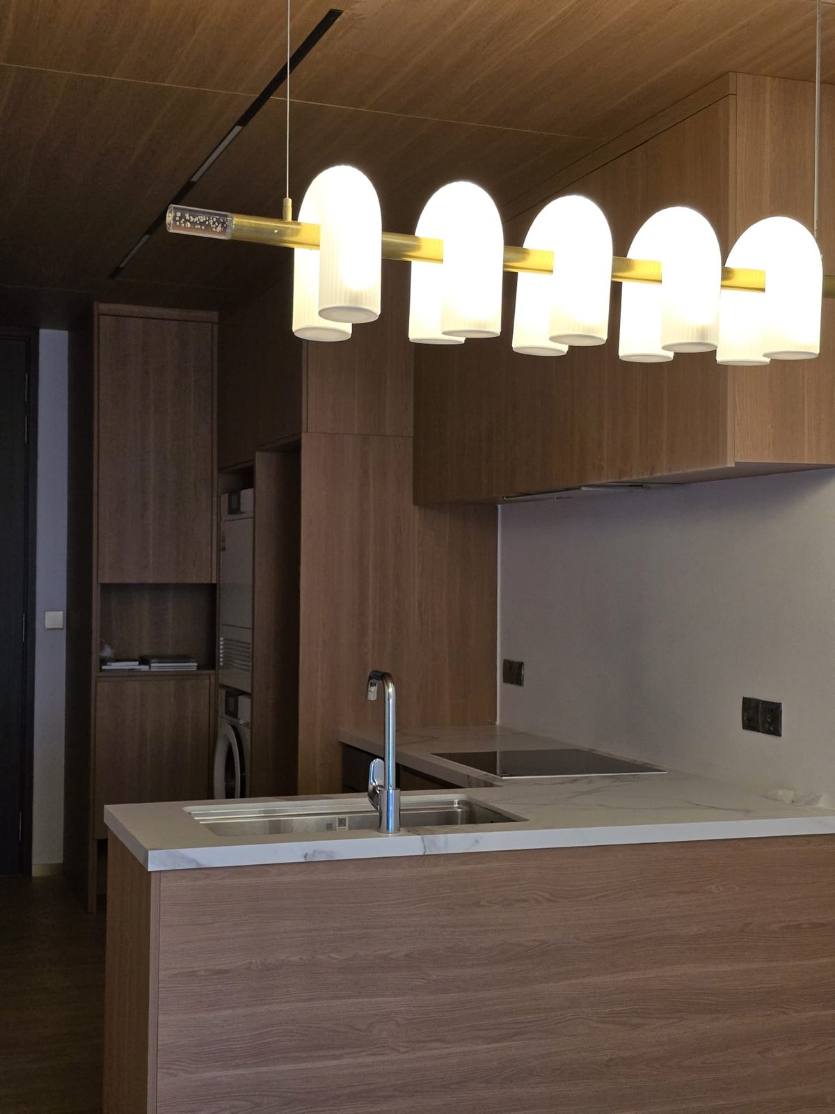 The kitchen with the ribbed-shade pendant lit against darker timber-toned joinery.