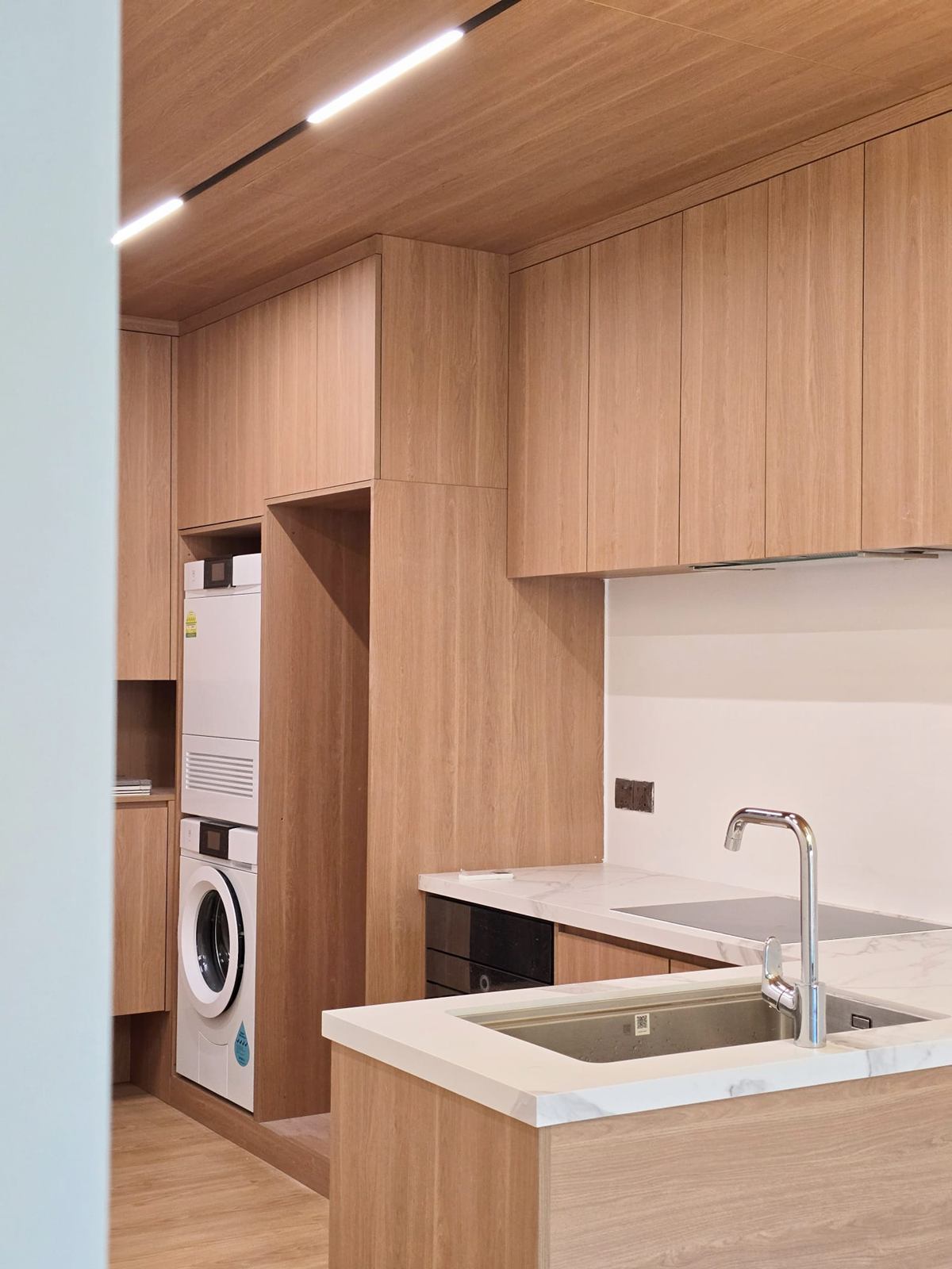 Timber-toned kitchen cabinets with a stacked washer and dryer built into a tall unit, and a white worktop in the foreground.